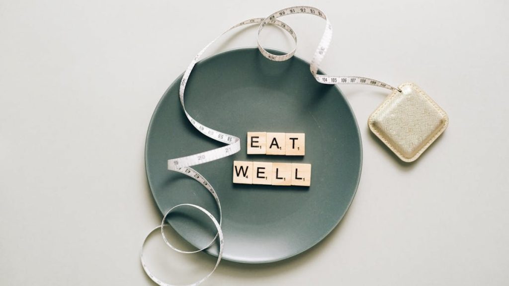 Flat lay of scrabble tiles spelling 'eat well' on a plate with a tape measure, promoting healthy eating.