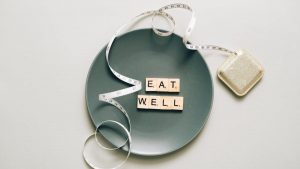 Flat lay of scrabble tiles spelling 'eat well' on a plate with a tape measure, promoting healthy eating.